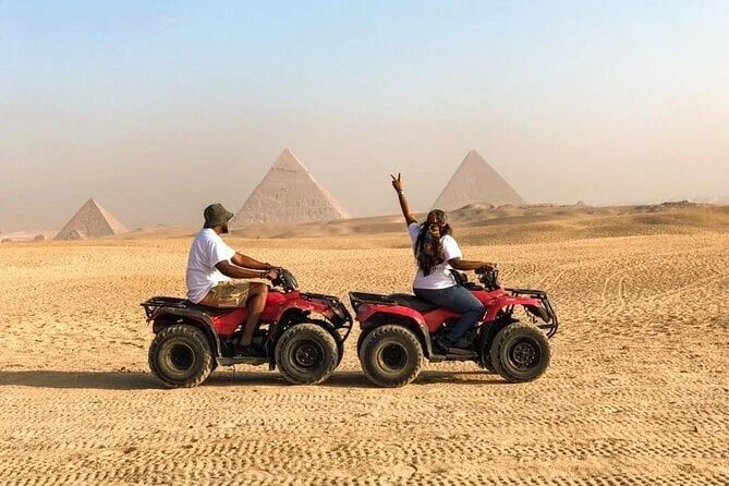 Two riders on red quad bikes cross the open desert, one raising a peace sign, with two pyramids visible behind them.