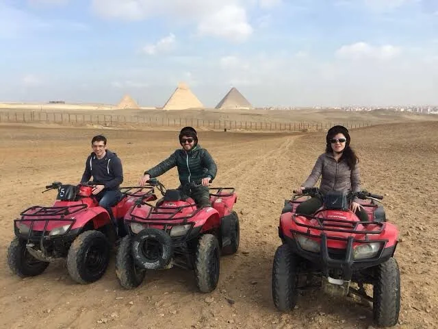 Three riders sit on red quad bikes posing for a photo with several Giza pyramids lined up in the background.