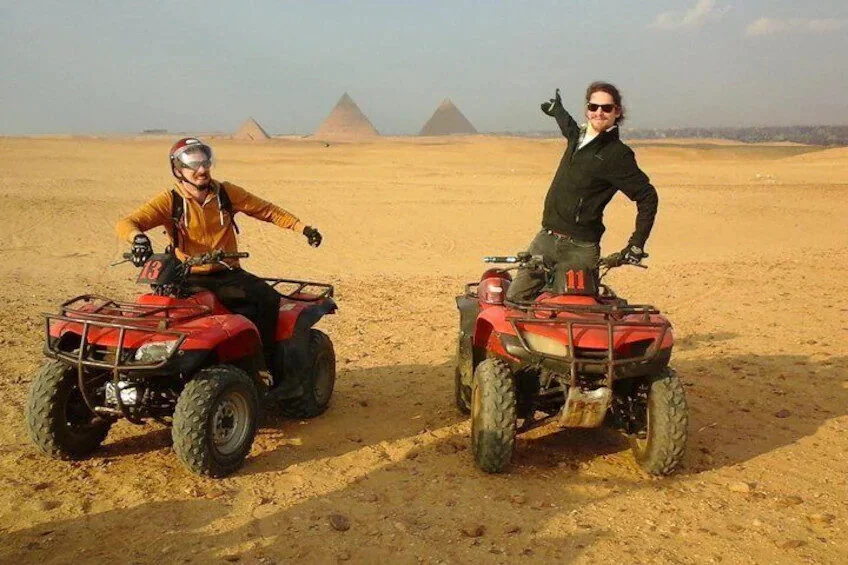 Two men pose on quad bikes in the desert, one giving a thumbs up, with two pyramids visible in the hazy distance.