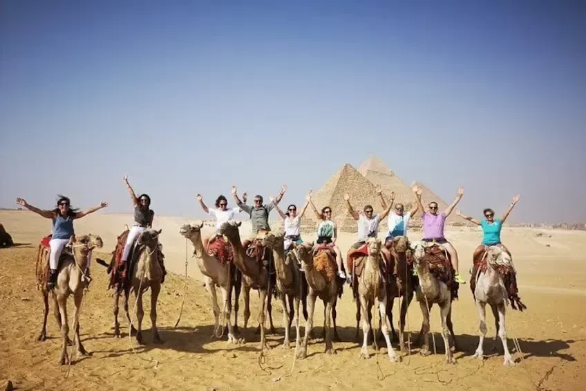 A line of ten tourists ride camels with arms raised in celebration, two pyramids visible in the background.