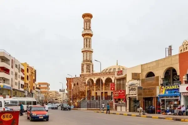 A downtown Hurghada street with a tall decorative minaret, shops and traffic passing by.