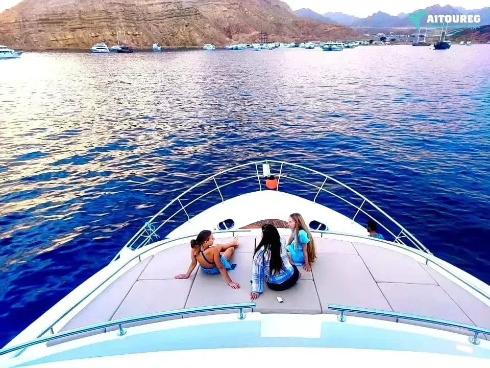 Three young women sitting and chatting on the front deck of a yacht at sunset, with anchored boats and mountains behind.