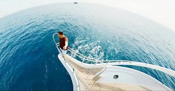 Fisheye view looking down from the yacht's bow at a man standing on the railing above the open sea.