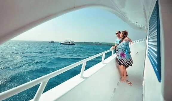 A couple standing arm in arm along the yacht's side railing, looking out at the sea and a passing boat.