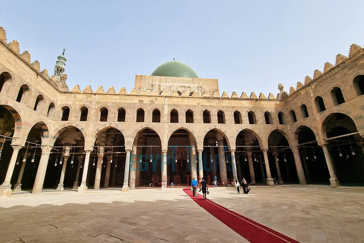 A stone courtyard surrounded by arched colonnades, with a green-domed structure and a red carpet leading across the floor.