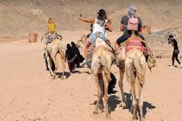 Tourists riding camels through the desert, one taking a selfie, with a guide on foot leading them.