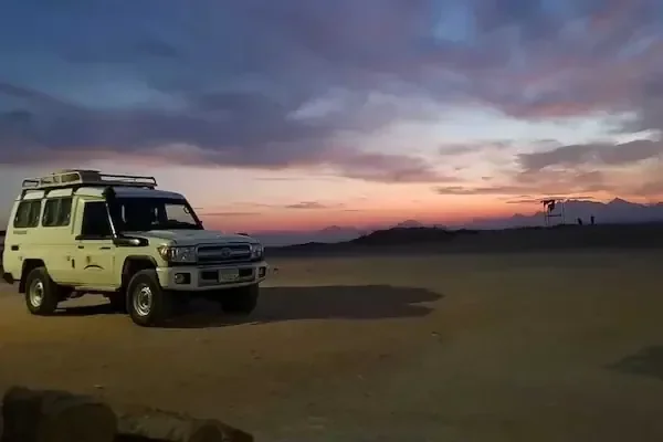A white 4x4 safari jeep parked in the desert at dusk under a colorful sunset sky.