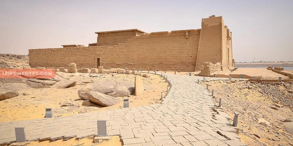A paved walkway leads toward the sandstone walls and pylon of an ancient temple on a rocky desert shoreline.