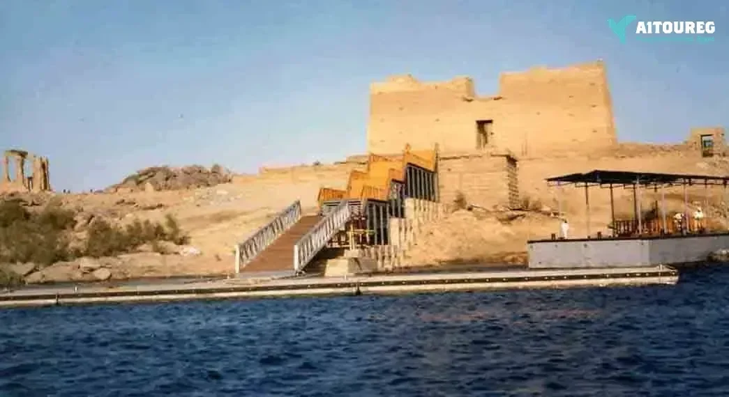 A wooden staircase and boat dock lead up to a sandstone temple on the shore of a reservoir, with another temple visible across the water.