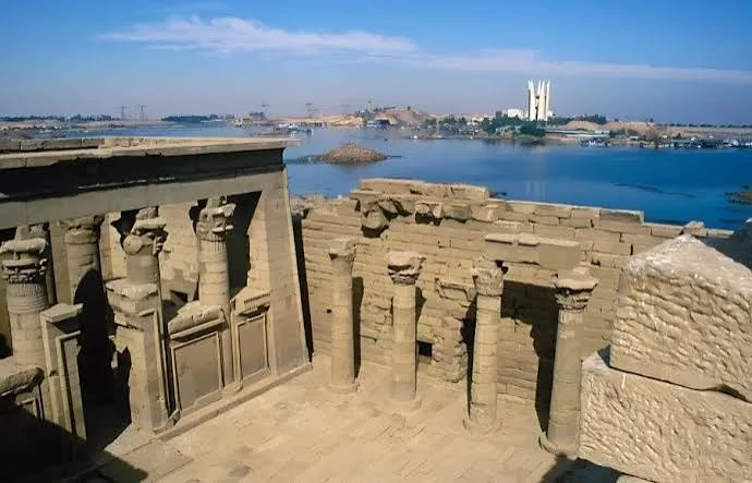 Ancient stone columns in the foreground with the sandstone temple and the waters of Lake Nasser beyond, a modern monument visible on the far shore.