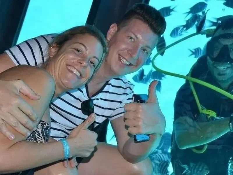 Two friends give a thumbs up inside the submarine cabin as a diver and fish are visible through the window behind them.
