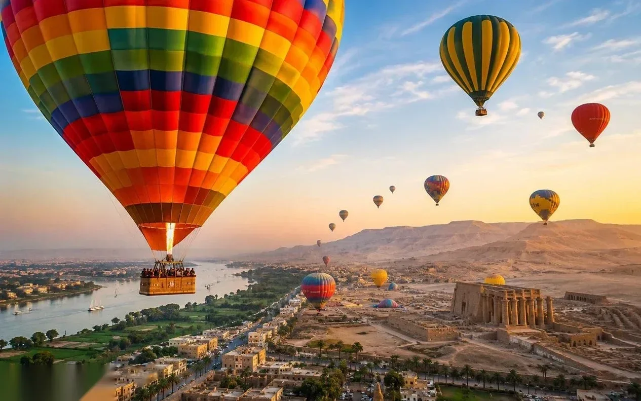 A large rainbow-striped hot air balloon flies over the Nile River and Luxor's temple ruins, with several other balloons in the sky.