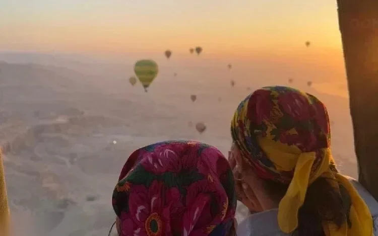 Two women wearing floral headscarves look out from a hot air balloon basket at other balloons scattered across the sunrise sky.
