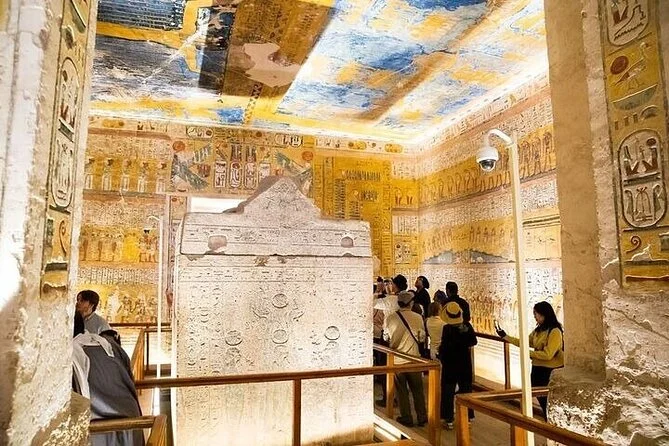 Brightly painted hieroglyphic reliefs in blue, yellow, and white cover the walls and ceiling of a royal tomb chamber, with visitors examining a stone sarcophagus.