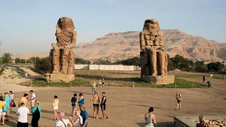 The two seated Colossi of Memnon statues rise from the desert plain with the Theban hills in the background, visitors walking below them.