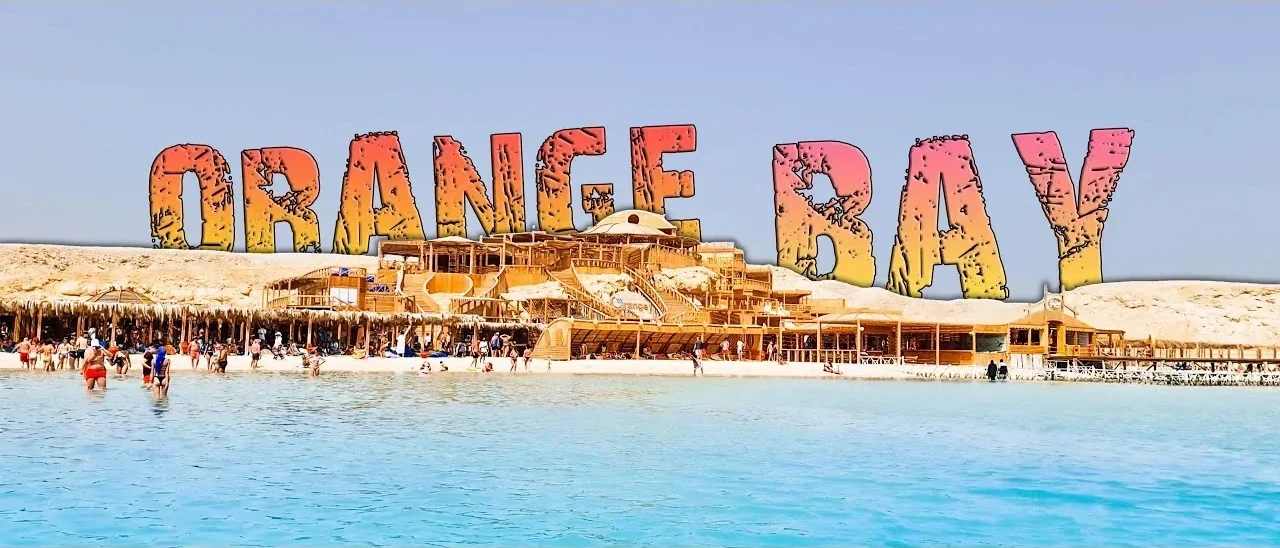The ORANGE BAY sign towers over a wooden resort complex, seen from the water with swimmers near the shore.
