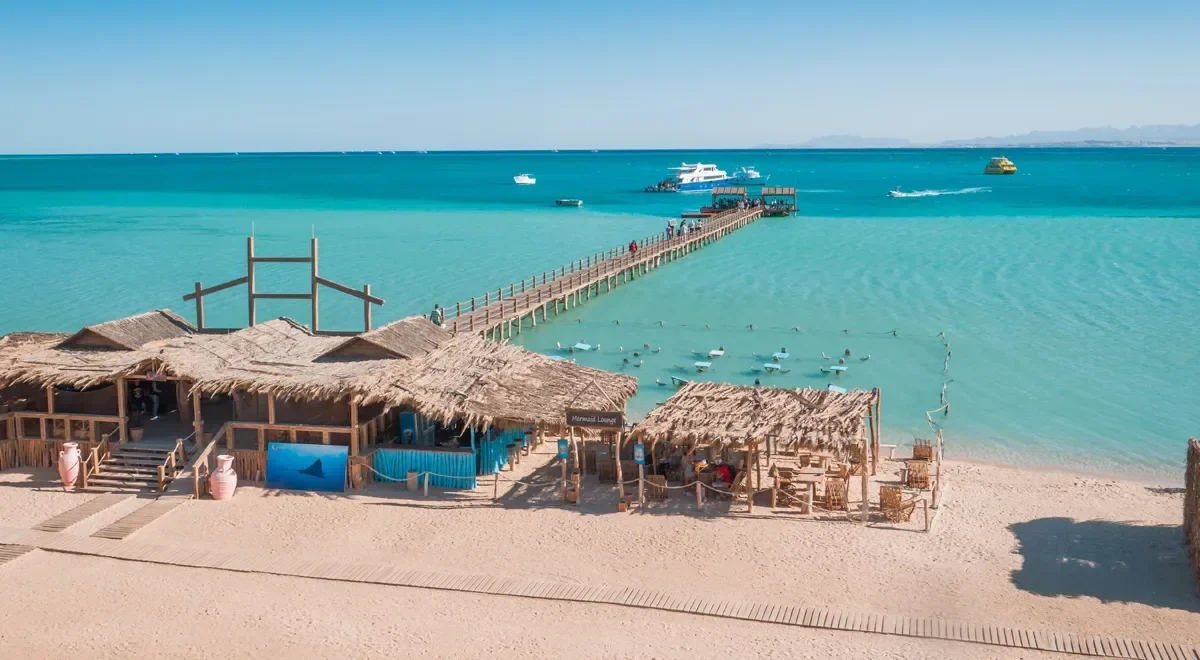 Aerial view of a wooden pier crossing turquoise water to a straw-roofed beach lounge, with boats anchored beyond.