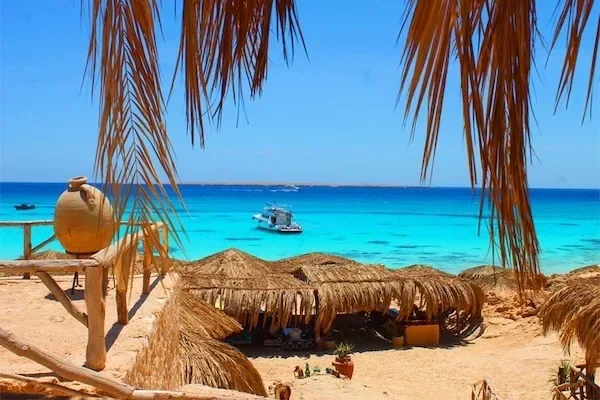 View through palm-frond shade of a moored motorboat and straw beach cabanas along the turquoise shoreline.
