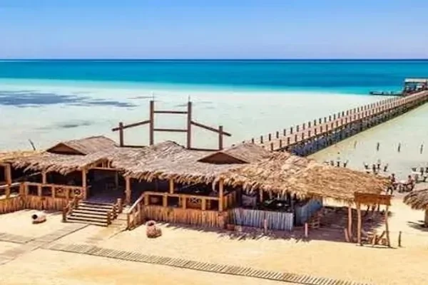 Cluster of thatched-roof beach buildings on the sand with a long wooden jetty stretching out over the turquoise sea.