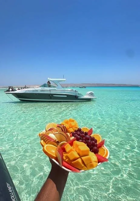 A hand holds a plate of fresh melon, orange, grape and apple slices with a small motor yacht anchored in shallow turquoise water behind.