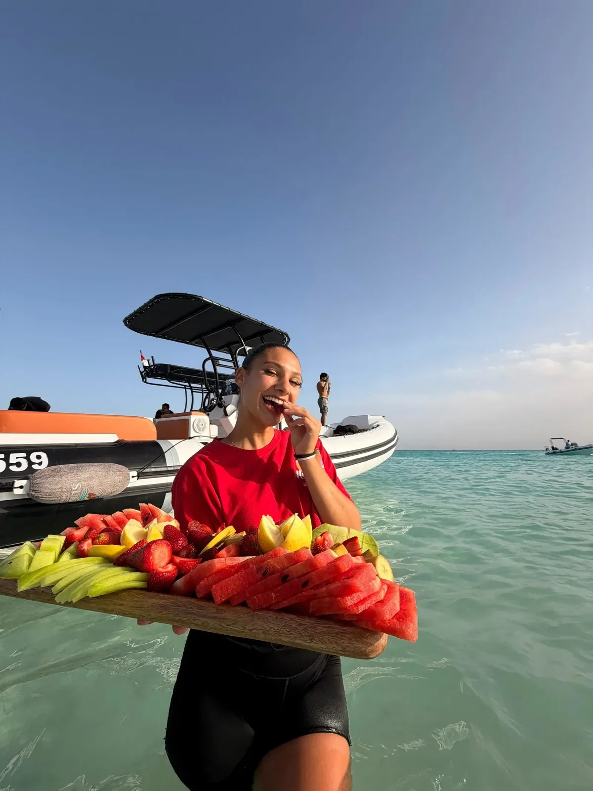 A woman holds a wooden platter of watermelon, cantaloupe and strawberries at the shoreline, smiling as she eats a slice, with a speedboat behind her.