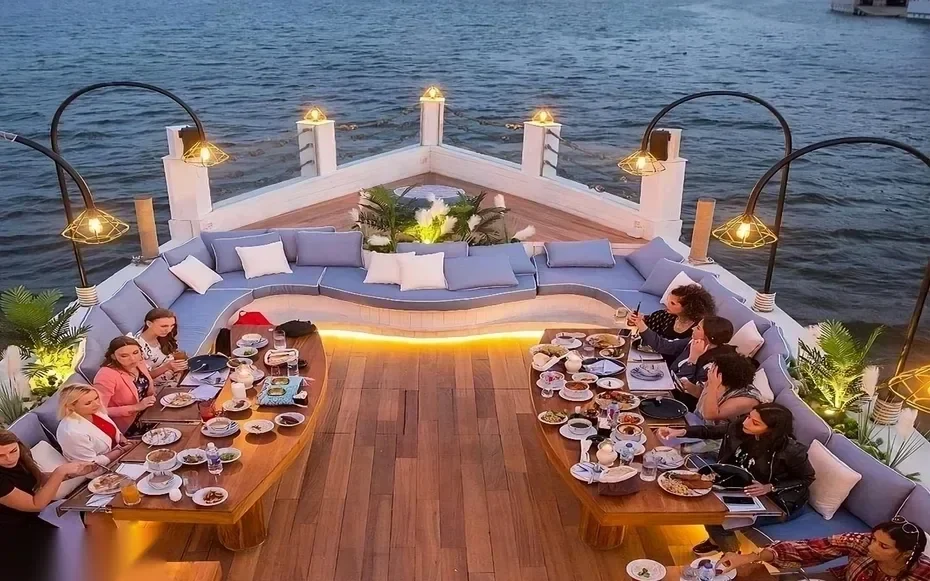 Aerial view of guests seated at curved sofa tables on the boat's bow, dining as evening lights come on over the river.