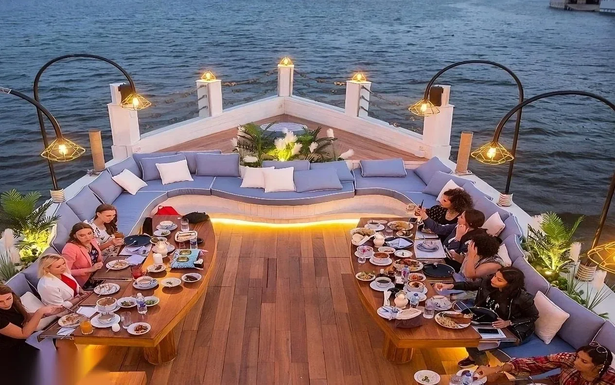 A similar aerial view of the same bow dining area, with guests eating and river lights reflecting on the water.