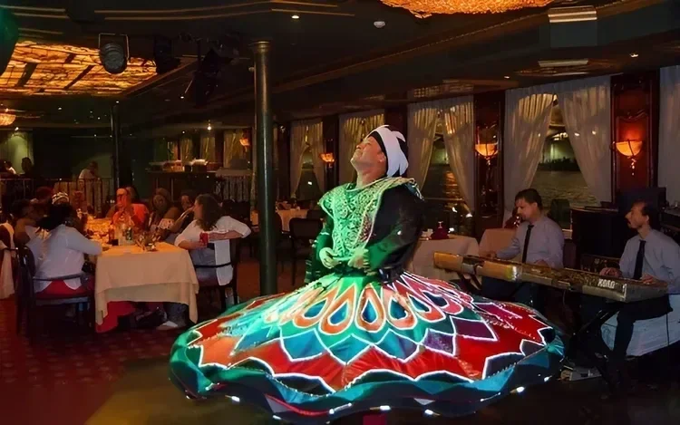 A whirling tanoura dancer in a green, red, and blue skirt performs mid-spin in the dining hall as diners watch.
