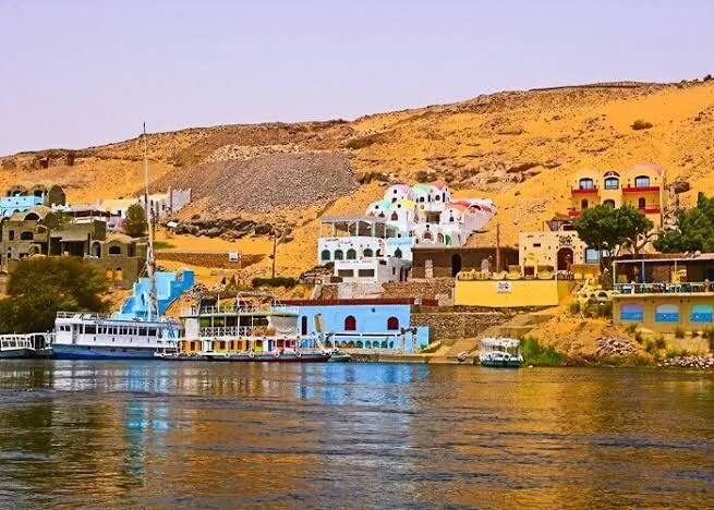 A cluster of tall Nubian houses in blue, yellow, and pink stacked up a rocky slope above the Nile, with boats moored below.