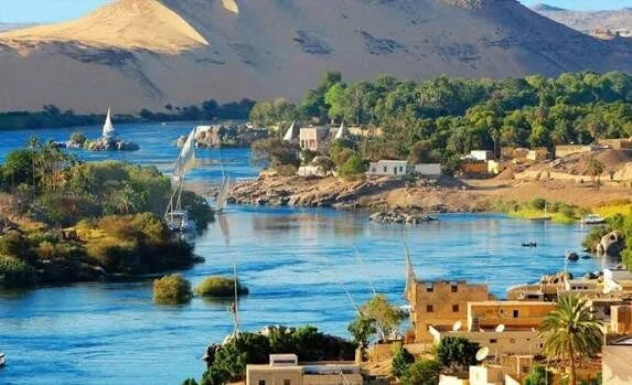 A Nubian village of domed and painted houses sits at the base of golden desert hills along the Nile, with boats docked at the shore.