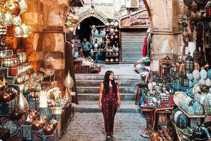 A woman in a patterned jumpsuit walks through a Khan el-Khalili alley lined with hanging lanterns, brass ornaments, and pottery stalls.