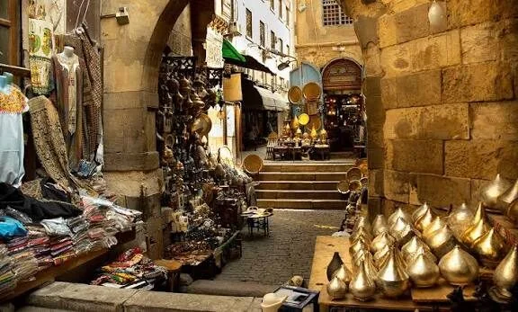 A market alley in Khan el-Khalili lined with hanging brass lanterns, textiles, and metalware stalls under a stone arch.