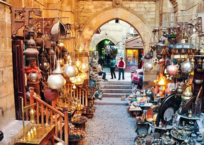 A narrow lantern-lined alley in Khan el-Khalili leads through a stone archway toward more market stalls, with a cat on the cobblestones.