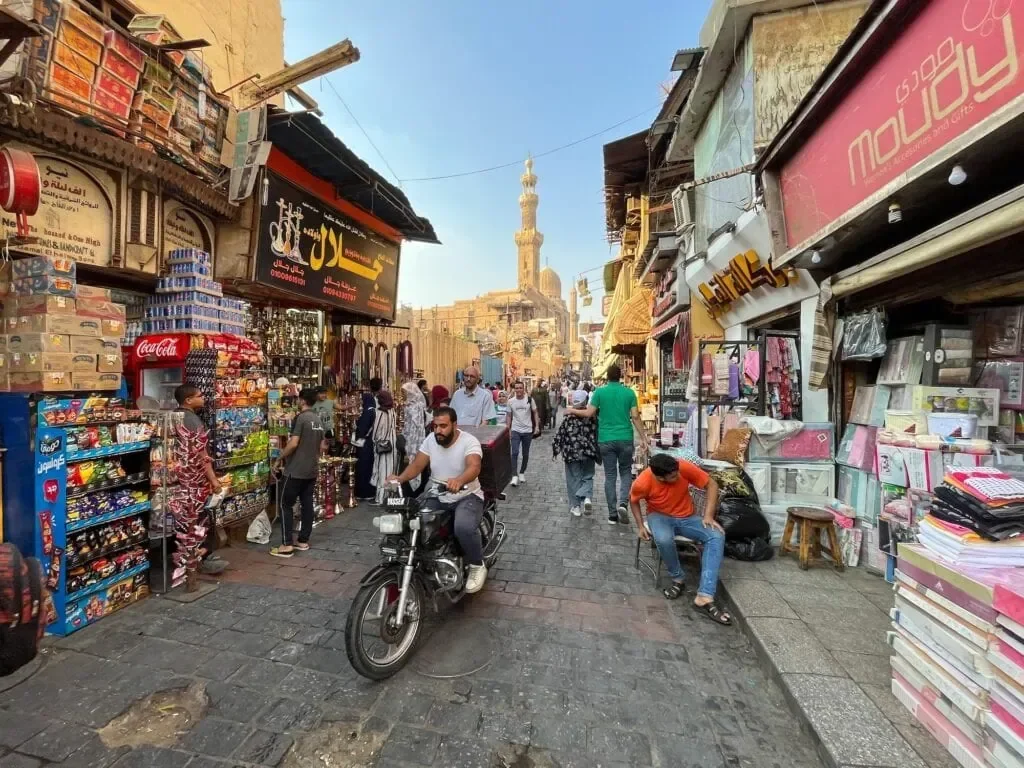 A busy Cairo market street lined with shops and stalls, with a motorbike rider and pedestrians passing beneath a distant minaret.