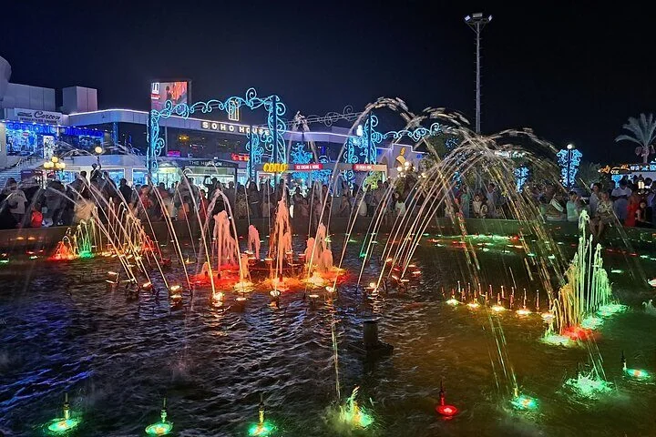 A choreographed fountain shoots dozens of illuminated water jets into the air at night, with crowds gathered around.