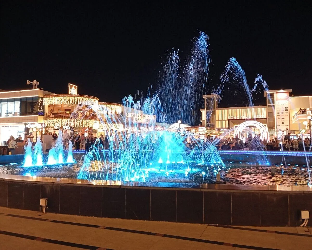 Tall blue-lit fountain jets spray upward in front of the Khan El Khalili Bazaar and Soho Mall storefronts at night.