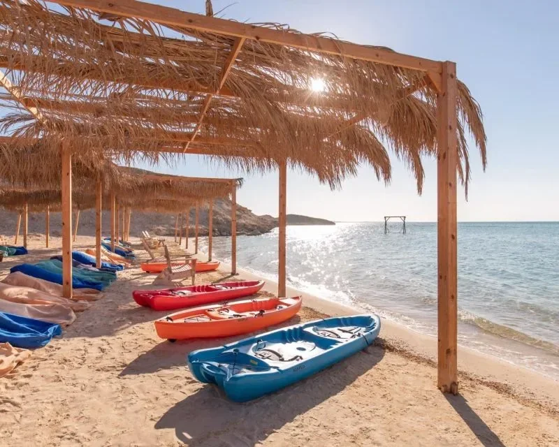 Orange, red, and blue kayaks lined up on the sand under wooden straw-roofed shelters at the water's edge.