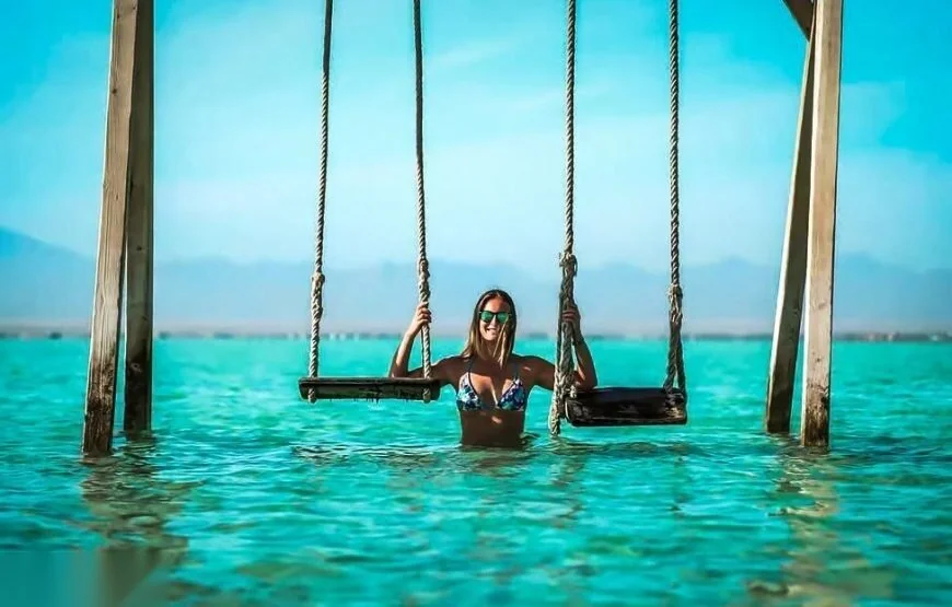A woman in a bikini stands between two rope swings partially submerged in shallow turquoise water, with hazy mountains in the background.