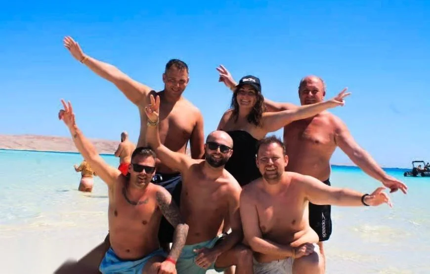 Six friends pose together in shallow water on the beach, making peace signs and smiling, with a beach buggy visible in the background.