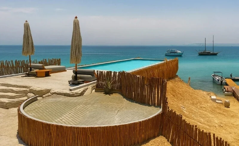 Aerial view of the Ozirea beach club's rooftop pool and wooden sun deck with straw umbrellas, overlooking anchored boats in a turquoise bay.