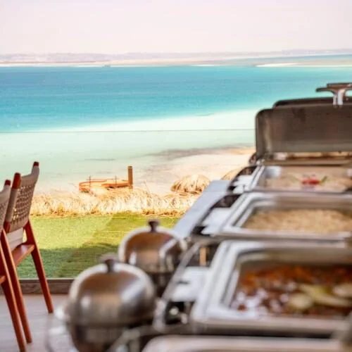 Rows of chafing dishes set up for a buffet lunch on an outdoor terrace overlooking a turquoise lagoon and white sandbar.