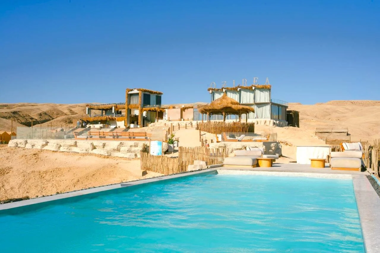 Wide desert view of the Ozirea beach club complex with a pool, thatched-roof buildings and a sign reading OZIREA against sandy hills and blue sky.