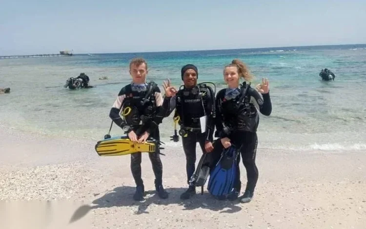 Three certified divers standing on a sandy beach in full gear giving OK hand signals, an instructor in a blue polo shirt between two students.