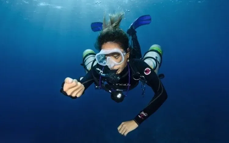Underwater portrait of a diver mid-descent with fins pointed upward, sunlight streaming down from the surface above.