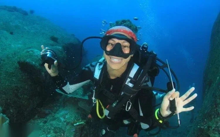 Close-up underwater portrait of a smiling diver on a rocky reef, holding a dive light and making a hand signal.