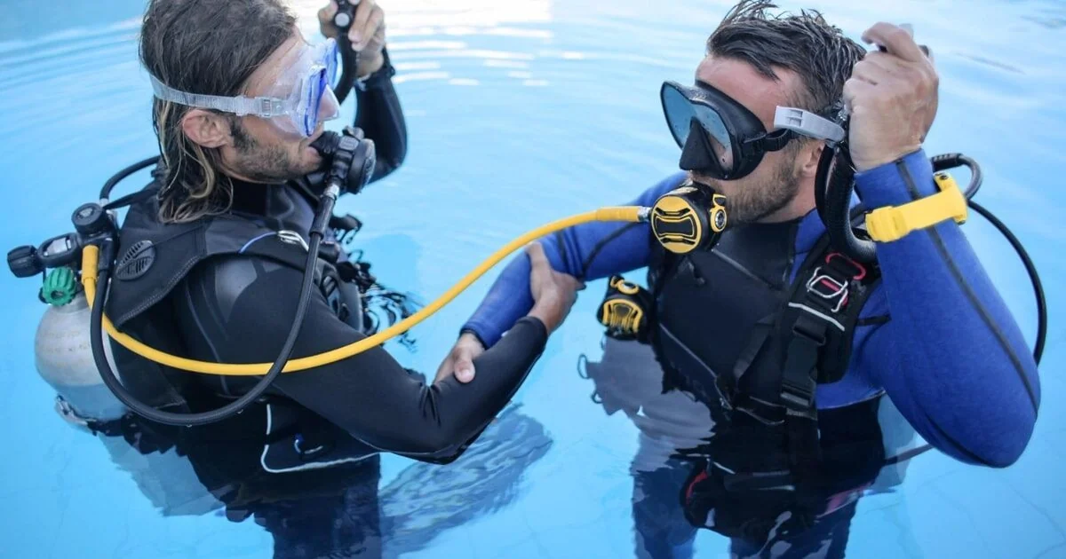 Two divers standing waist-deep in a swimming pool practicing an alternate-air-source exercise, connected by a yellow regulator hose.