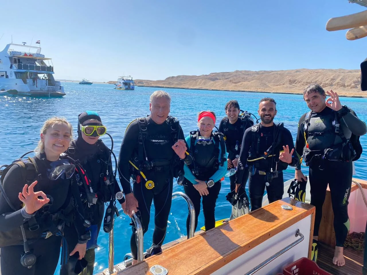 Group of seven divers posing on a dive boat's deck giving OK signs, with the desert coastline and other boats in the background.