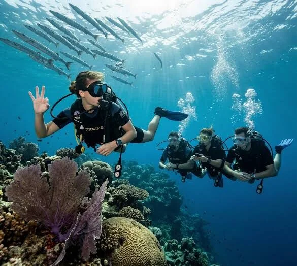 Divemaster trainee giving an OK sign near a school of barracuda above a coral reef with a purple soft coral fan, three students following behind.