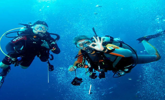 Two divers giving OK signs while mid-water, one holding an underwater camera, bubbles rising around them.