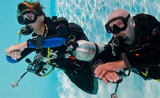 An instructor and student float horizontally side by side in a training pool, each wearing a single scuba cylinder.